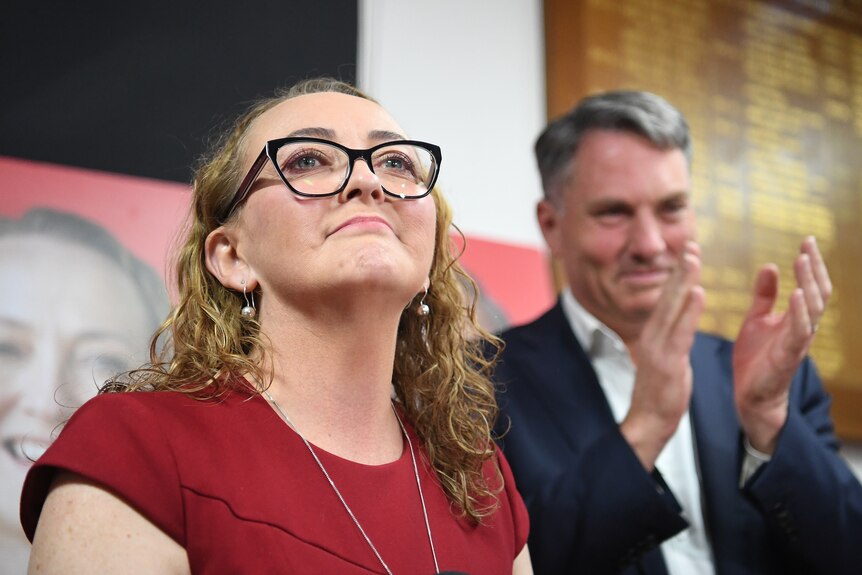 Mary Doyle smiles in a red top, with Richard Marles clapping out of focus behind her.