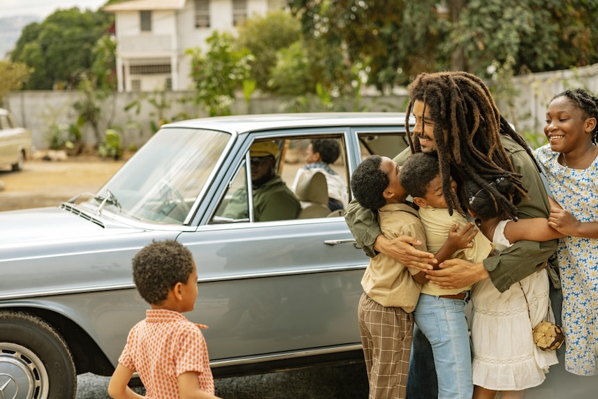 A man standing in front of a car with children, some of whom he has in an embrace