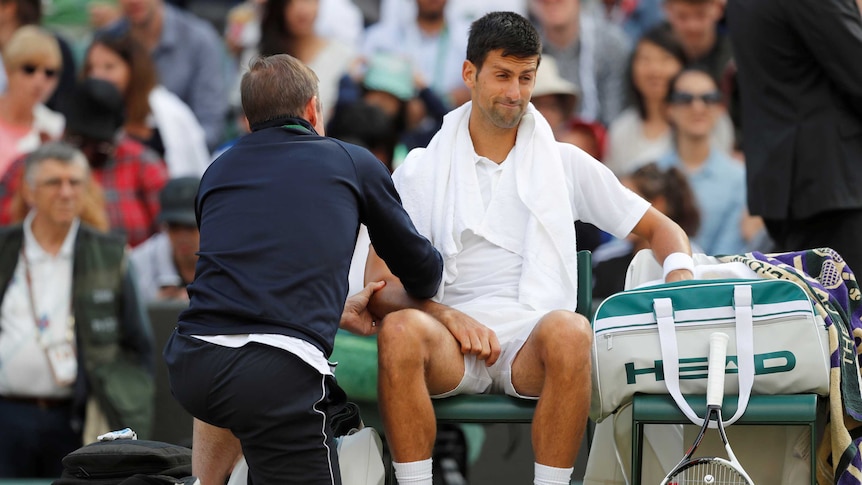 Novak Djokoivc seated as a trainer gives him medical treatment for a right elbow injury.