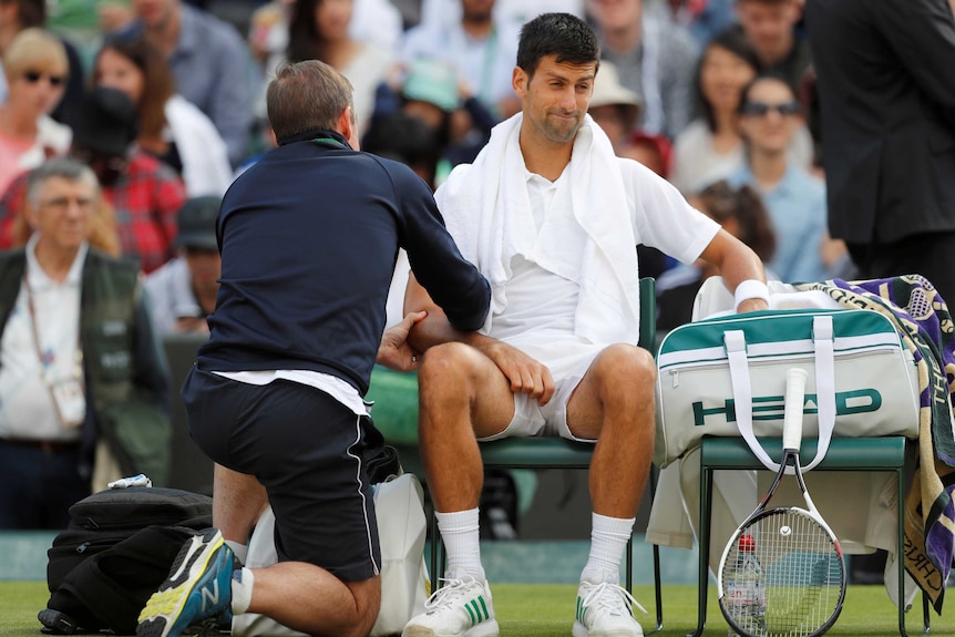 Novak Djokoivc seated as a trainer gives him medical treatment for a right elbow injury.