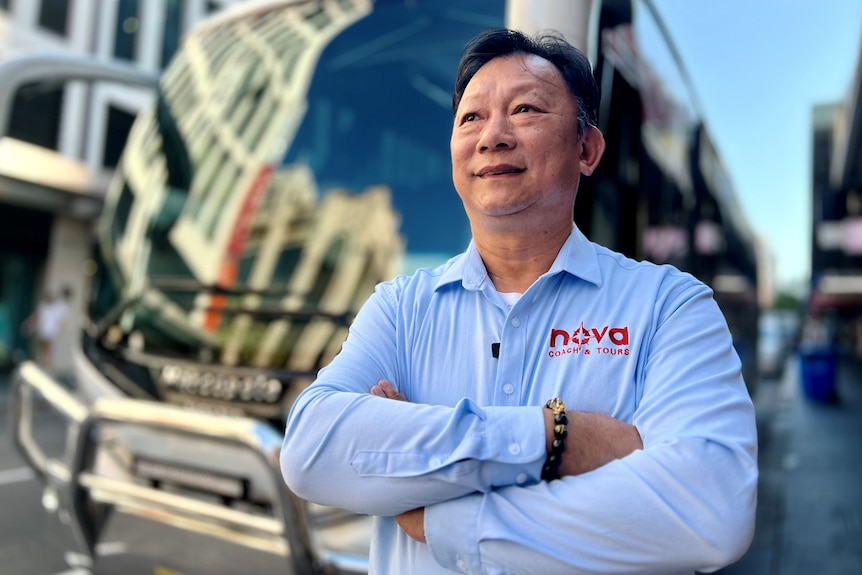 A man wearing a uniform stands in front of a bus, crossing his arms and smiling.