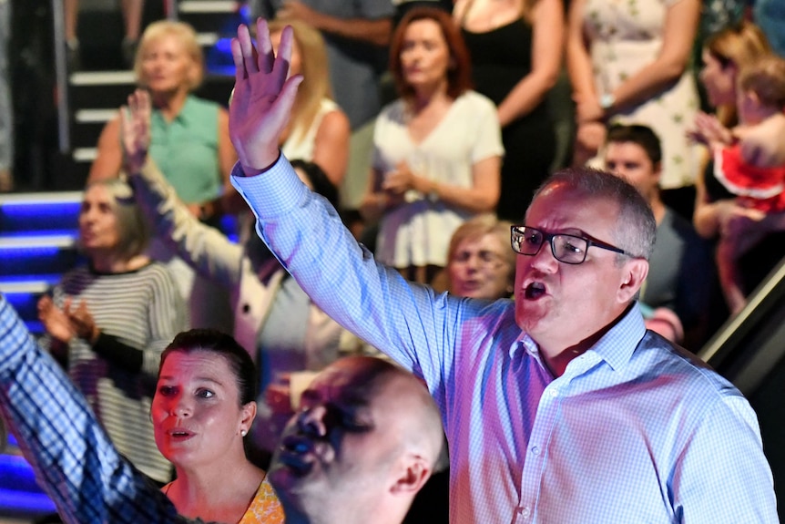 Scott Morrison raises one arm up towards the sky while singing in a crowded church, a man in front follows suit with closed eyes