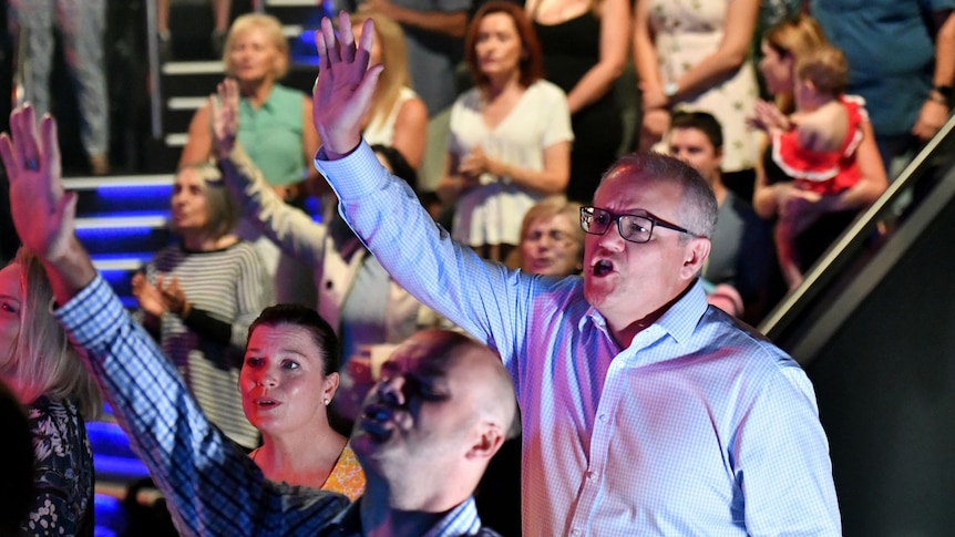 Scott Morrison raises one arm up towards the sky while singing in a crowded church, a man in front follows suit with closed eyes