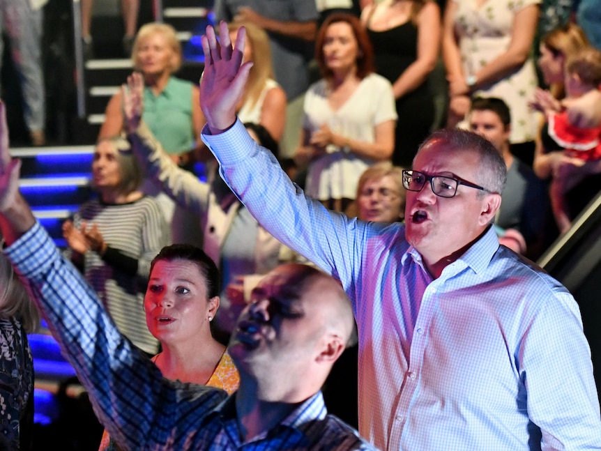 Scott Morrison raises one arm up towards the sky while singing in a crowded church, a man in front follows suit with closed eyes