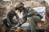 Two men in military uniforms kneel over an old, dirty bomb. One of the men has a tool in his hand.