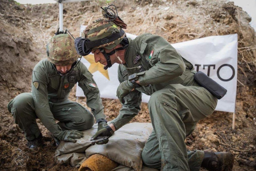 Two men in military uniforms kneel over an old, dirty bomb. One of the men has a tool in his hand.