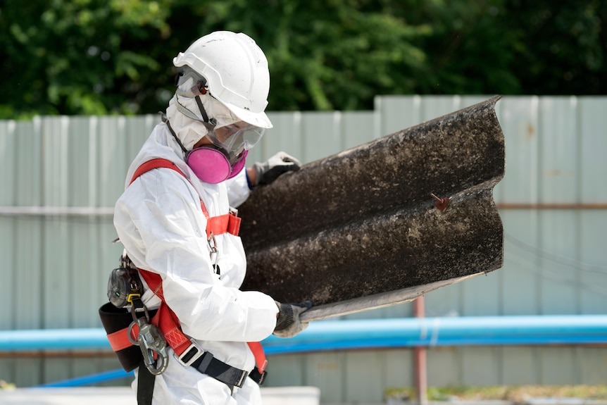 Person in white protective bodysuit with hard hat, mask and glasses holds material surrounded by construction site.