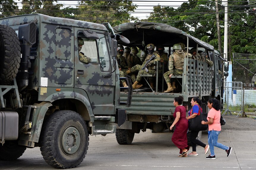 People in uniform sit in the back of a truck as civillians cross the road.