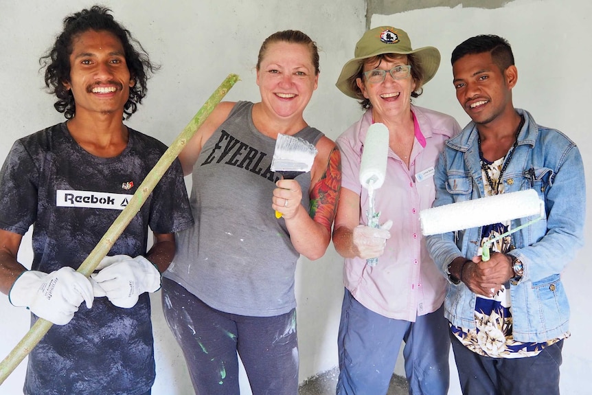 Two men and two women hold used paint brushes with white paint on them they are all smiling