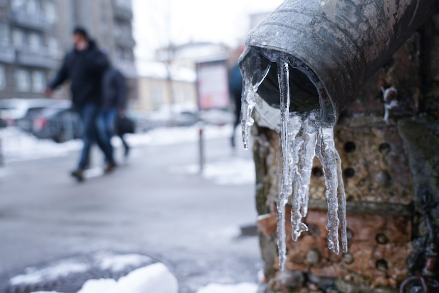 A man on a street walks passed a pipe covered in little icicles.