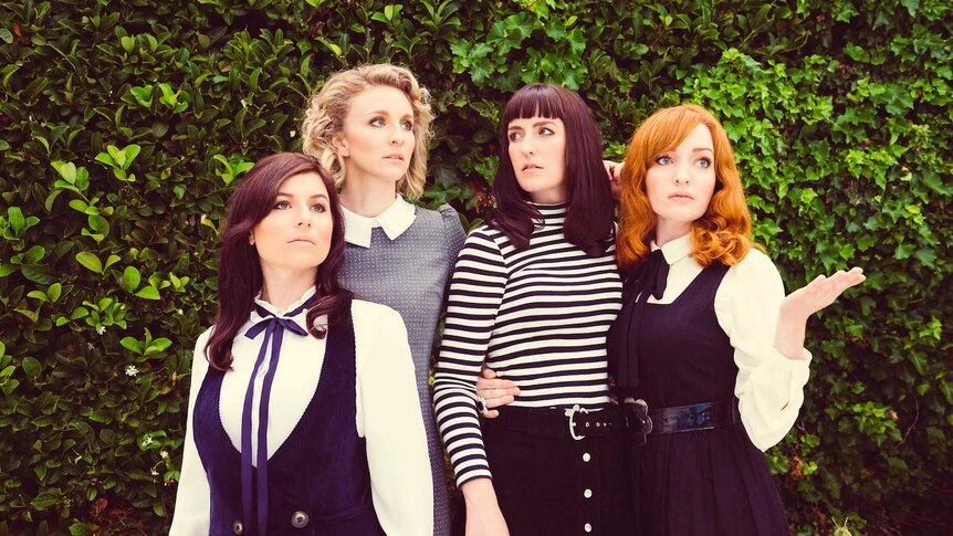 Four well-dressed female musicians stand in front of a lush green hedge