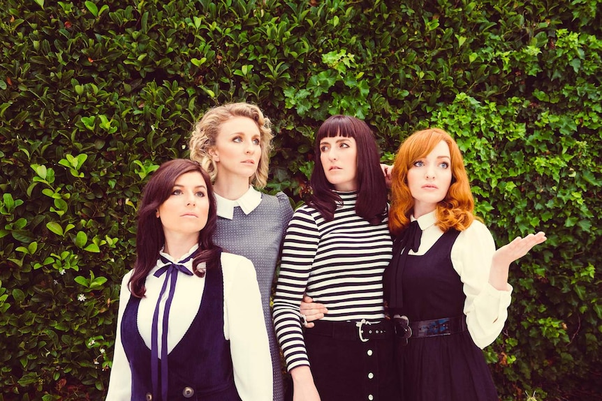 Four well-dressed female musicians stand in front of a lush green hedge