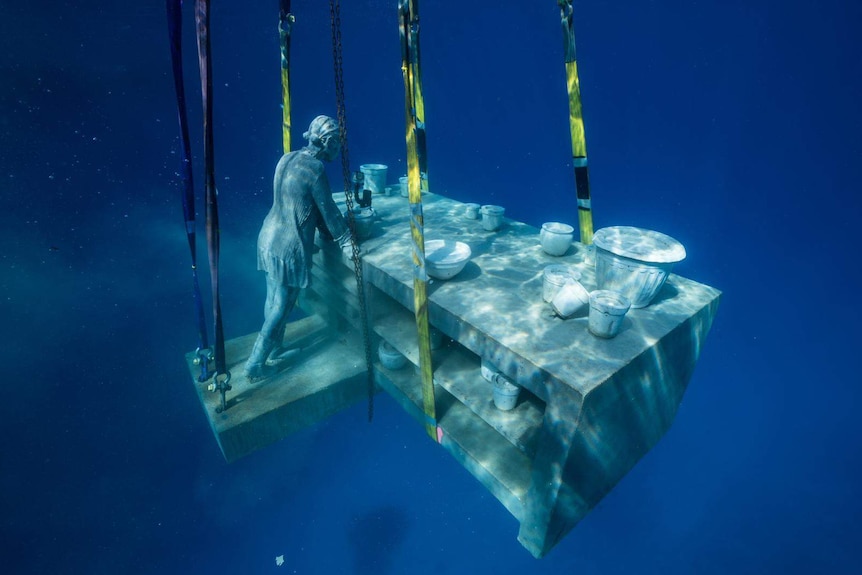 A sculpture of a woman standing at a bench with bowls on it is lowered down under the sea.