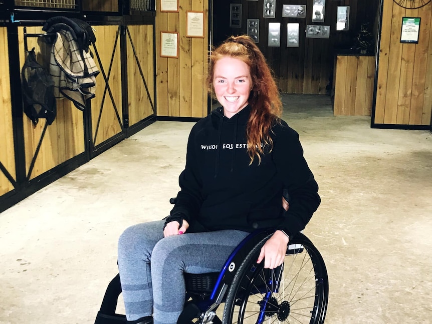 A young woman in a wheelchair is in a horse stable, she smiles at the camera.