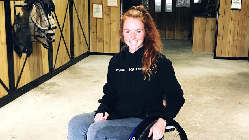 A young woman in a wheelchair is in a horse stable, she smiles at the camera.