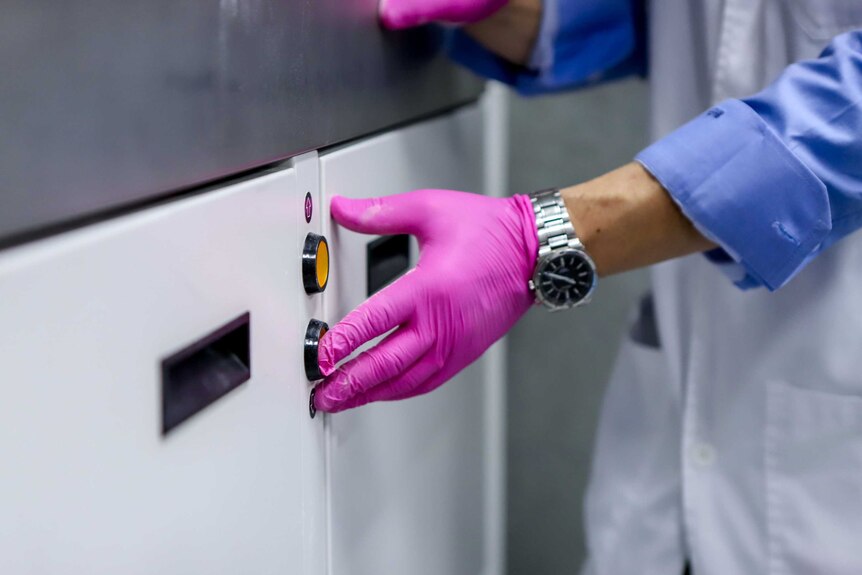 A pink-latex gloved-hand pushes a red button on a white and silver 3D printing machine