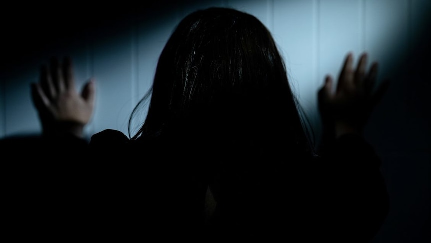 A dark haired woman pushing against a white wall with both hands. 