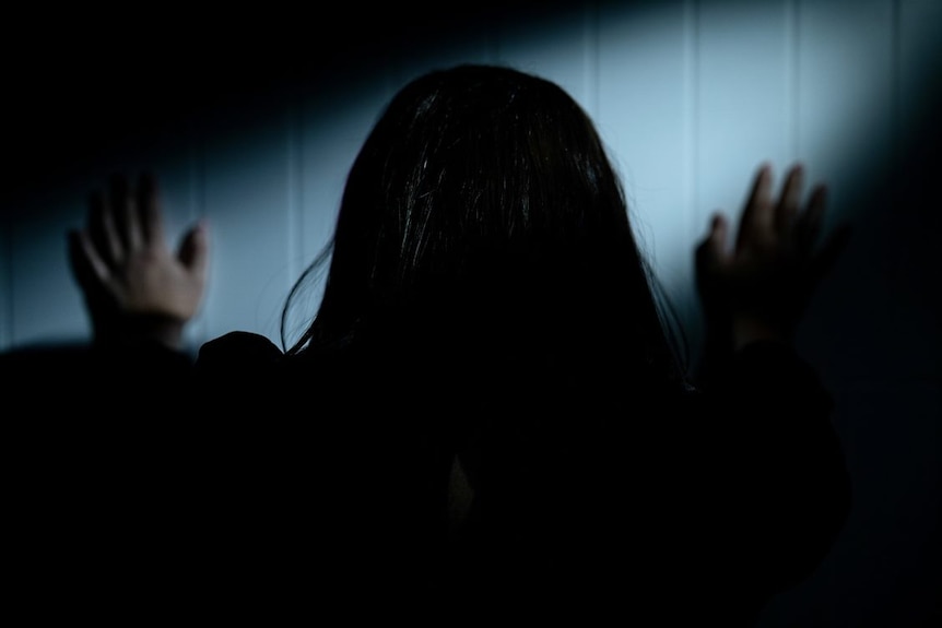 A dark haired woman pushing against a white wall with both hands. 