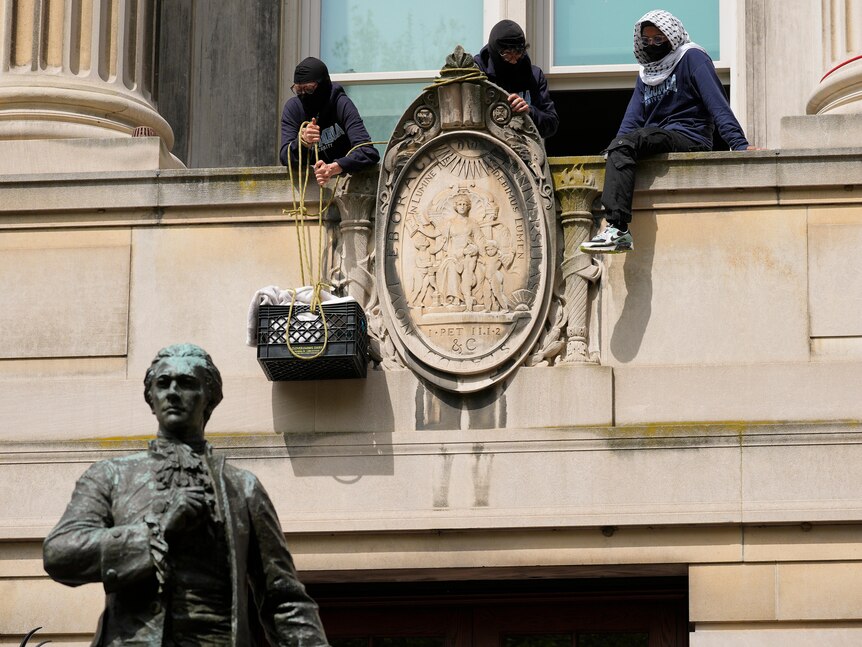 Three people wearing hoods sit on a balcony - one is hoisting up a black basket full of goods