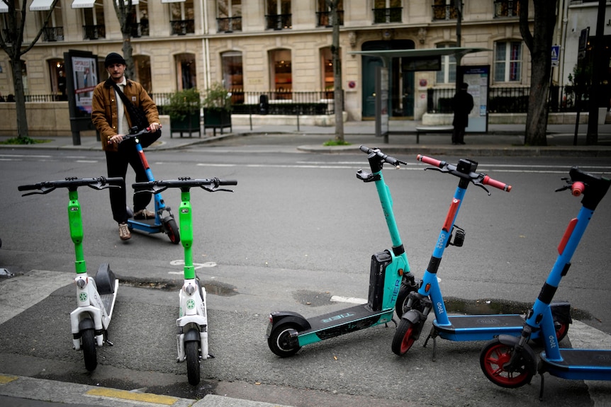 A man standing on an electric scooter looks at a group of five scooteres painted in green-and-white, pale blue and turquoise.
