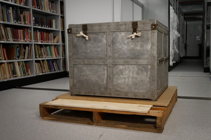 A large steel trunk on a wooden pallet with bookshelves in the background