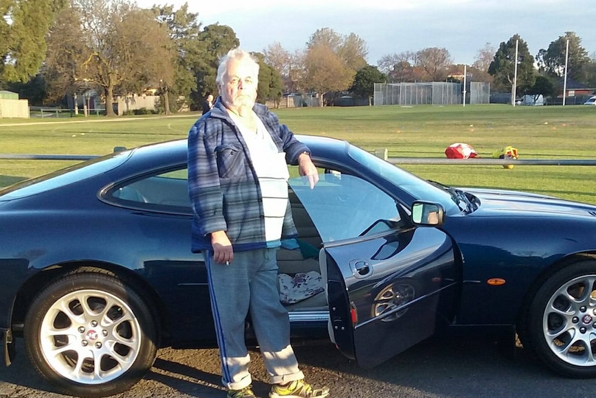 An elderly man stands next to a car, with the driver side door open