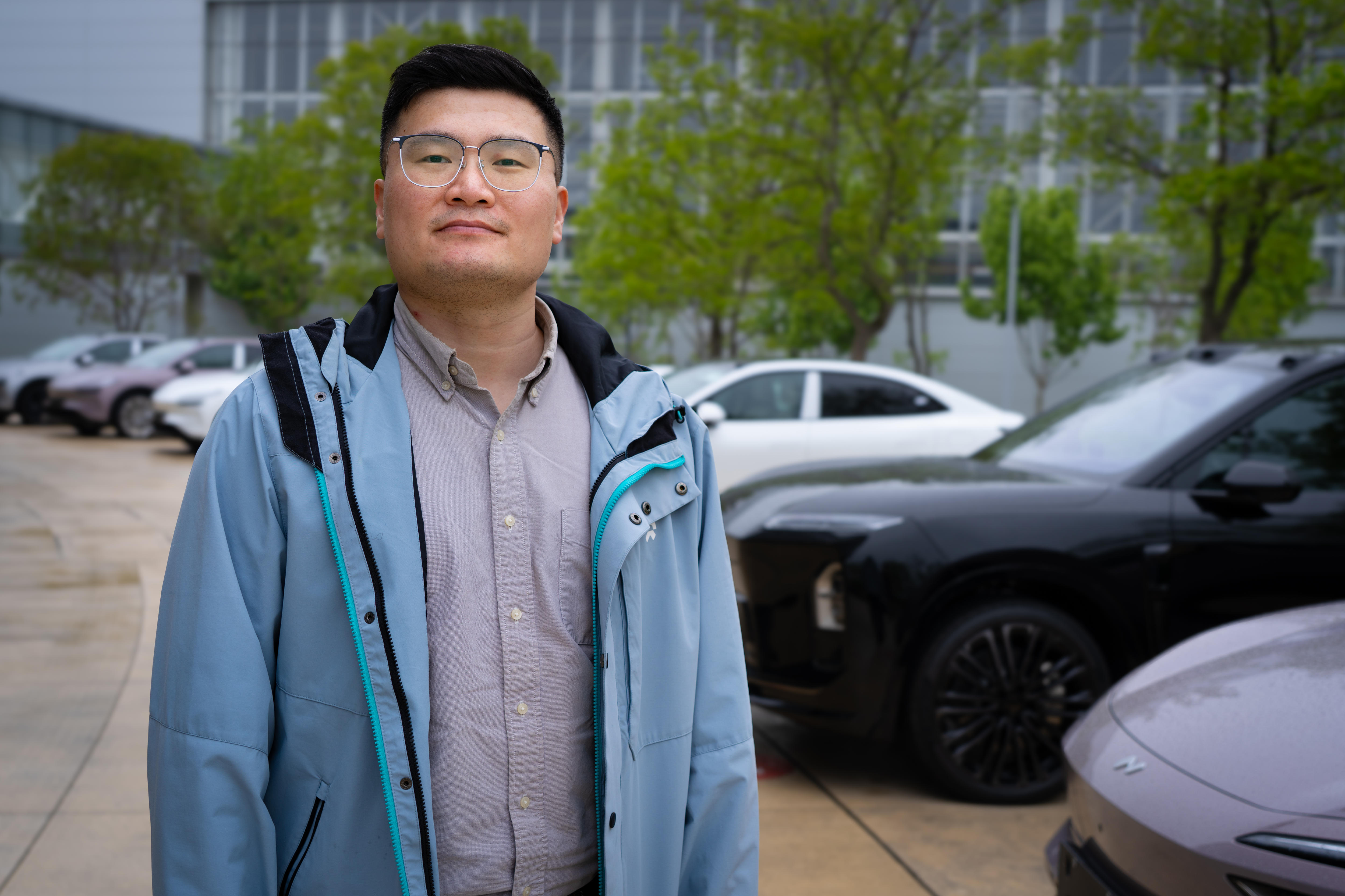 A man in a blue jacket and grey shirt, standing outside with a row of cars behind him.
