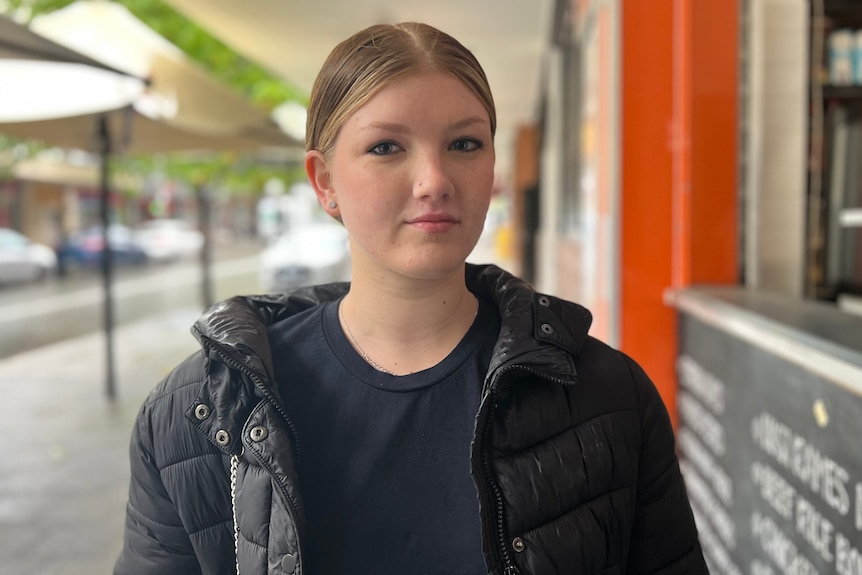 A head and shoulders shot of a young woman wearing a black coat and dark shirt standing on a street posing for a photo.