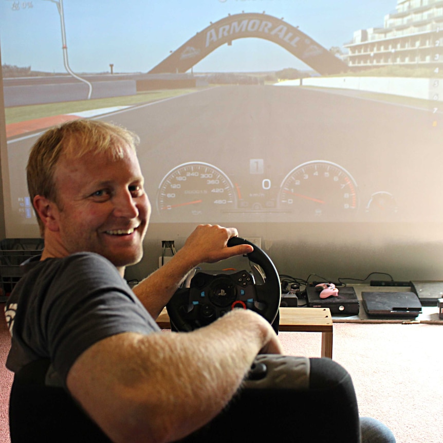 A man sits in a car seat in front of a projector screen with a car racing game.