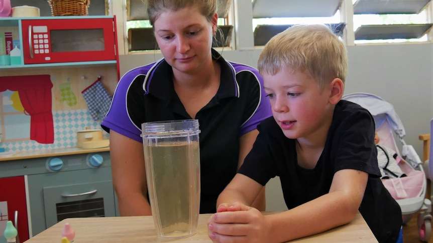 A blonde woman and blonde child sit at a kids table in a day care centre and look at a plastic cup of discoloured water.