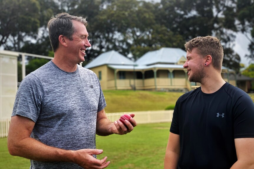 A man holding a cricket ball looks at his young adult son, laughing.