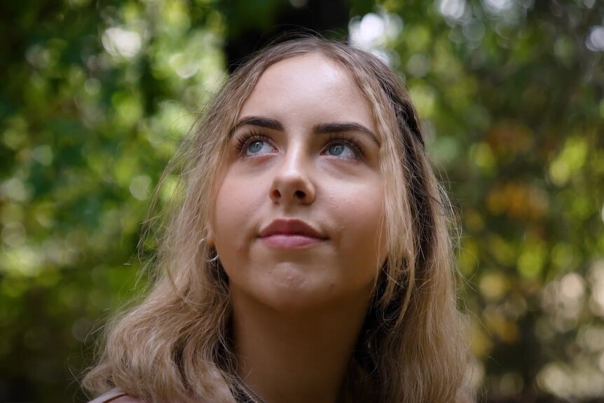 A young woman with blonde hair looks up, behind her are green trees