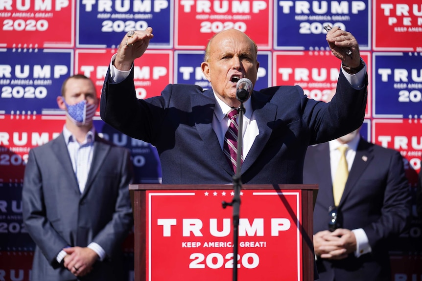 Rudy Giuliani holds both hands above his head while speaking. There are Trump 2020 banners around him