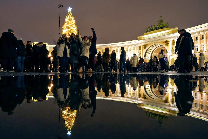 A group of people take a photo with a selfie stick in front of a lit-up Christmas tree.