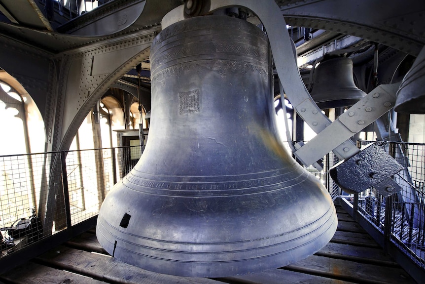 A large iron bell is pictured hanging amid a cast-iron structure located behind gothic windows.