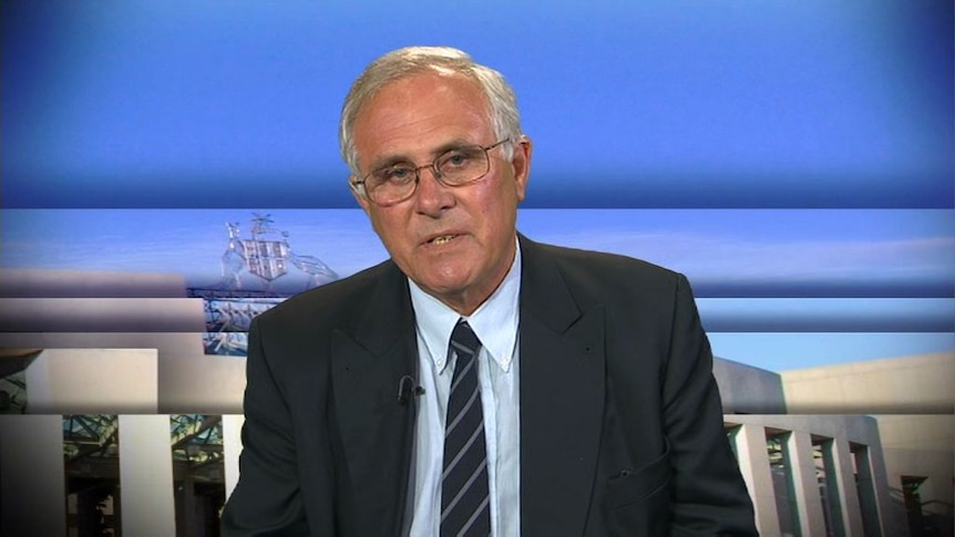 A man in a jacket, tie and glasses in a television studio.