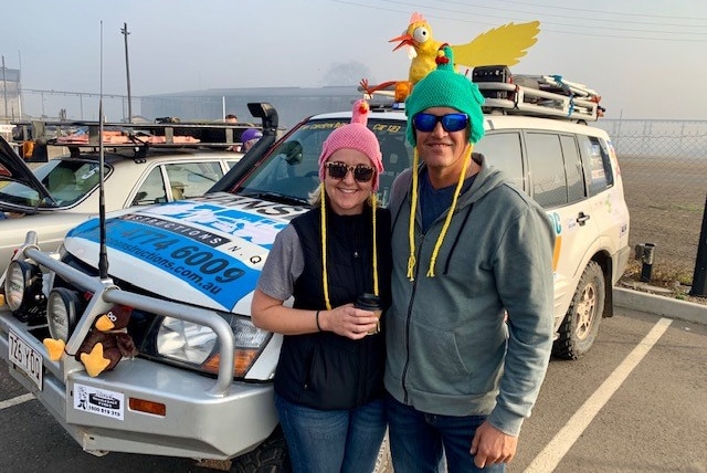 A man and woman wearing beanies shaped like chickens stand in front of their car with a fibre glass chicken on the roof