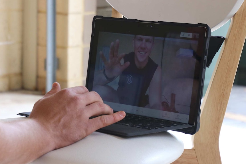 A laptop sits on a chair in a garage with a man on the screen waving and a hand on the keyboard.