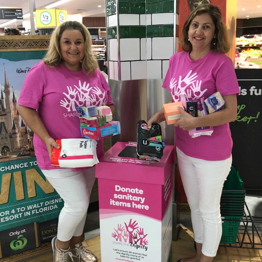 Two women in pink shirts holding sanitary items above a donation box