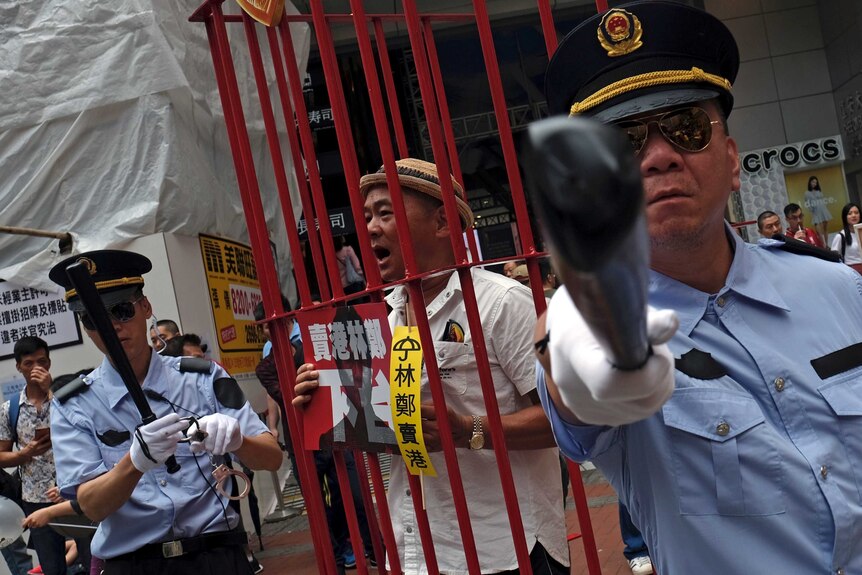 A protester dressed as a Chinese police officer points a fake baton at you as a man behind them is inside a red cage.