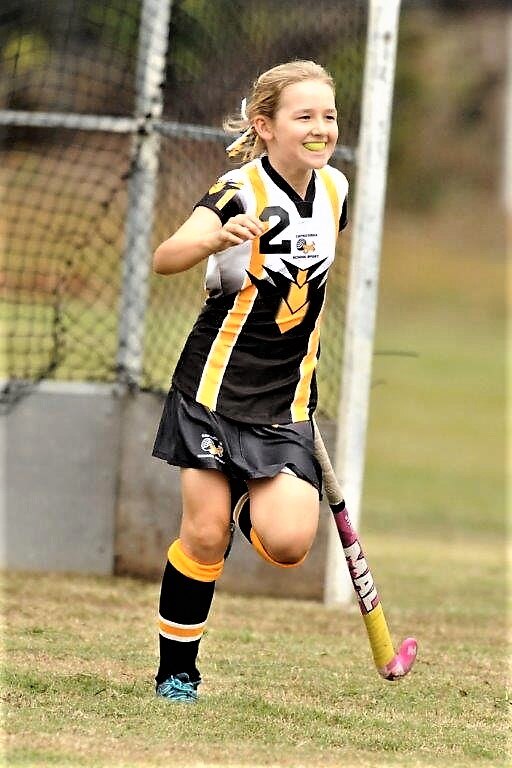 A young girl on a hockey field, with a black, white and yellow uniform, running and smiling.