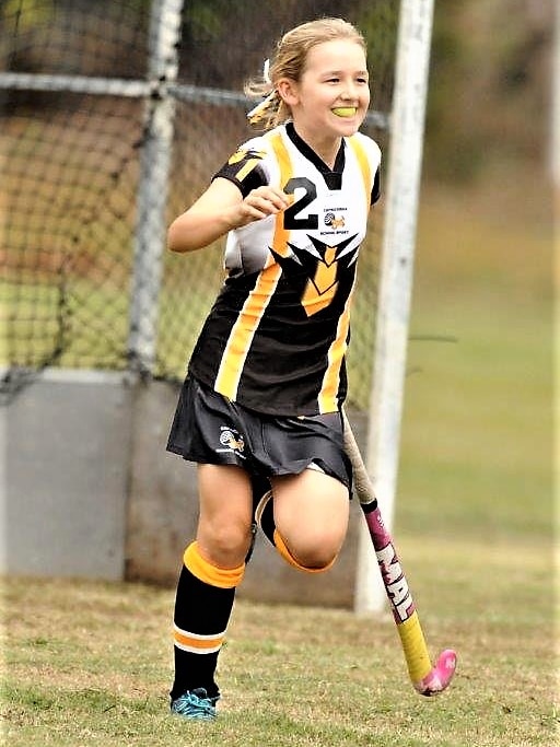 A young girl on a hockey field, with a black, white and yellow uniform, running and smiling.