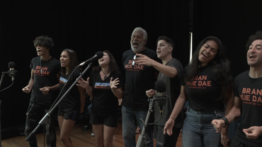 In front of microphones, Ernie Dingo (centre) and six other performers sing.