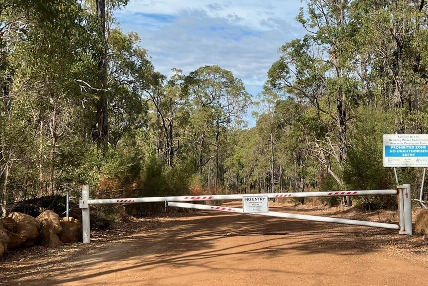A white boom gate with a sign saying no entry on a red dirt track with trees surrounding it