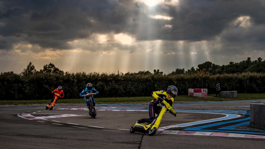 Three electric scooters racing on a track. The riders' outfits & scooters are brightly coloured: one red, one blue, one yellow
