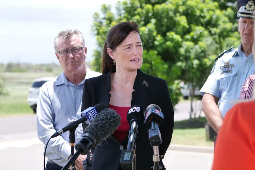 A woman with dark hair tied back stands at a microphone outside speaking to media