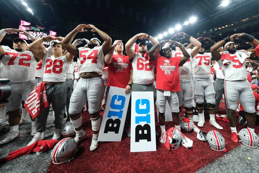 A group of American footballers stand in a group and hold their arms in an arc above their heads while looking out at the crowd