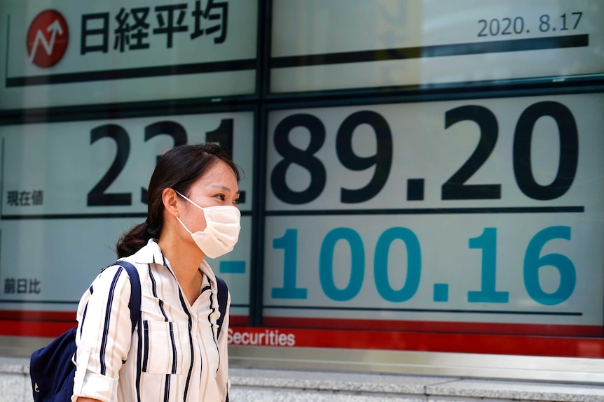 A woman wearing a face mask walks past a electronic screen showing numbers.