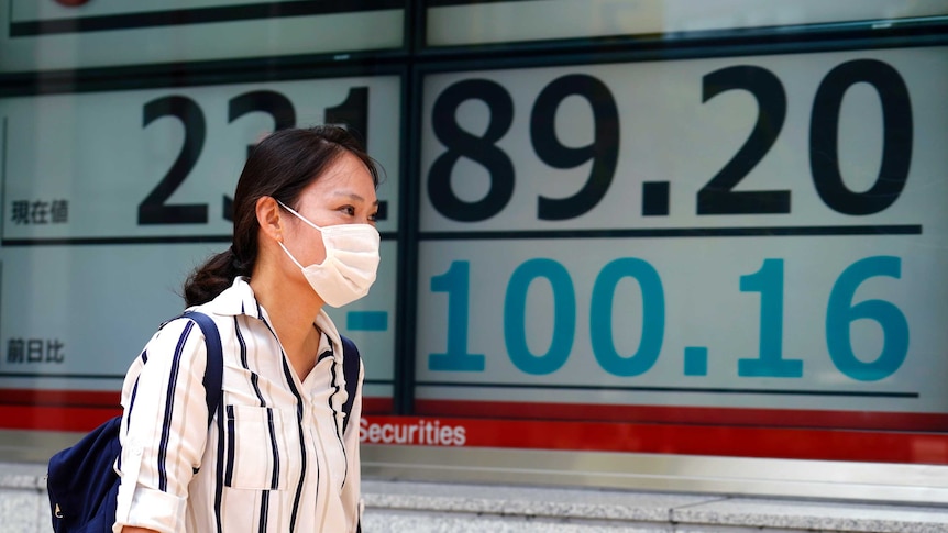 A woman wearing a face mask walks past a electronic screen showing numbers.