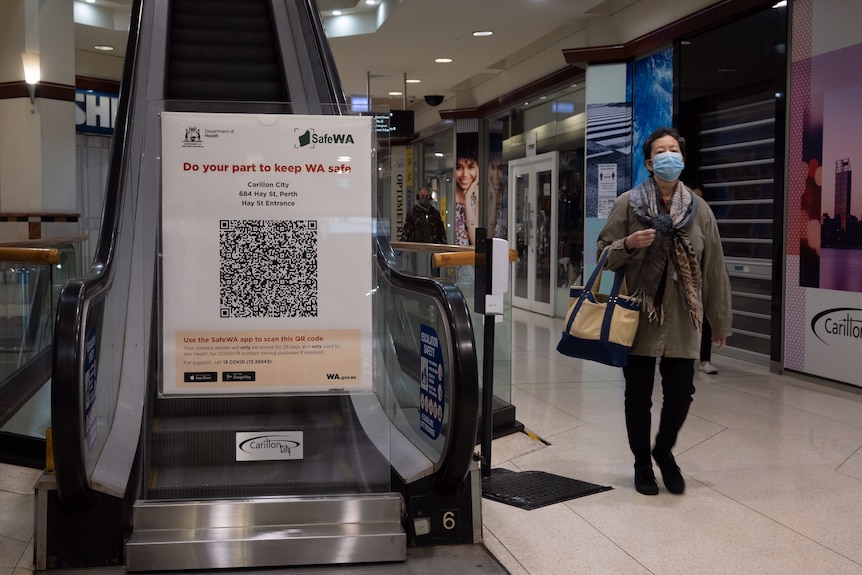 A woman wearing a mask walks past a QR code sign blocking off an escalator.